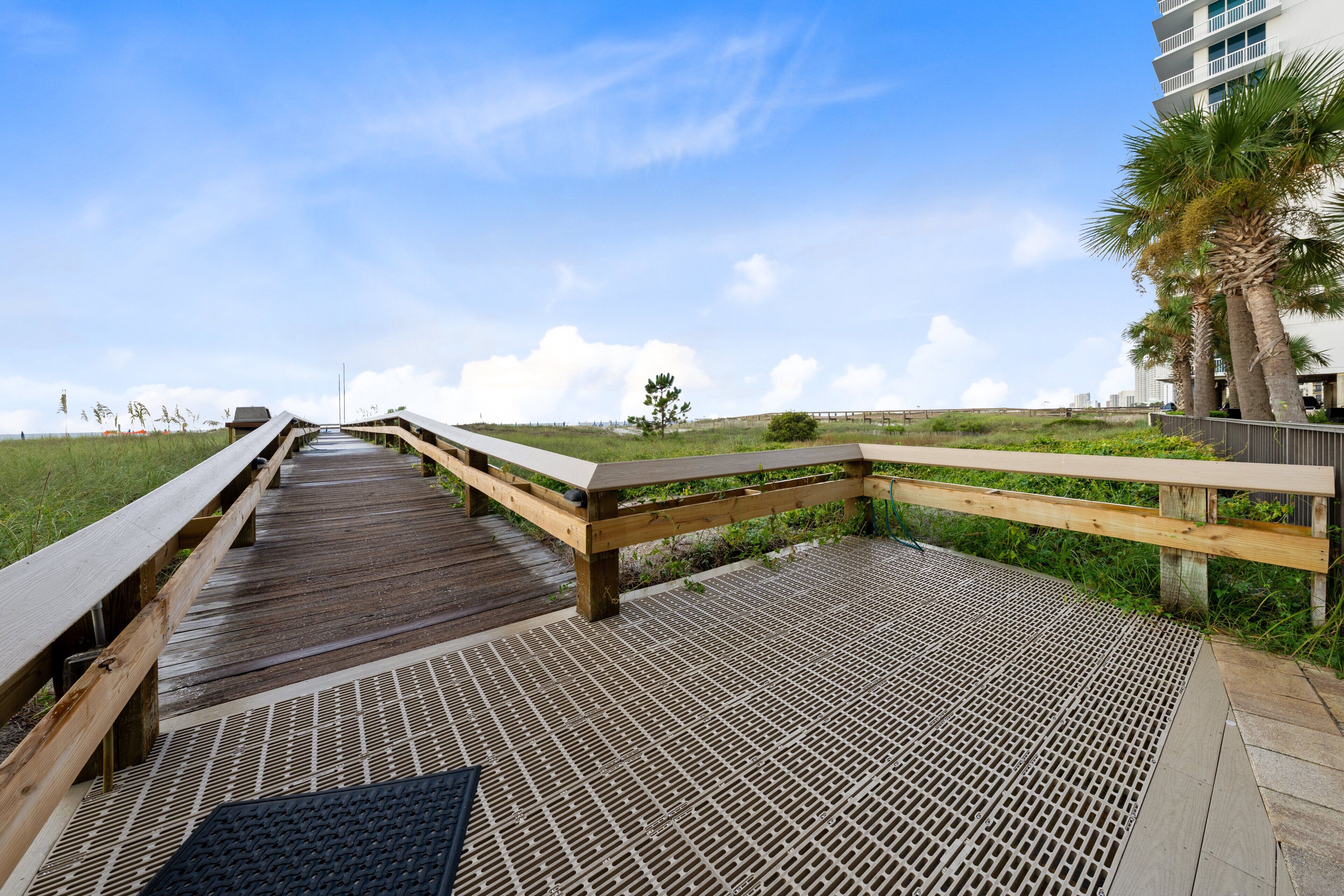 A wooden boardwalk leading to a beach, with a grassy area and palm trees on the right.