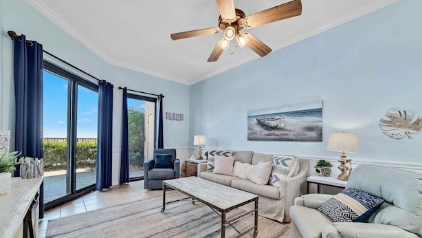 A spacious living room with a ceiling fan, a large window with curtains, a sofa, a coffee table, and a painting on the wall. - Phoenix East 009 - Ground-floor Gulf-front Condo - Sleeps 8 - Orange Beach, AL (Orange Beach)