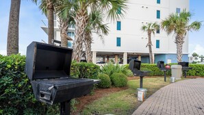 An outdoor grill area with a brick pathway, palm trees, and a building in the background. - Phoenix East 009 - Ground-floor Gulf-front Condo - Sleeps 8 - Orange Beach, AL (Orange Beach)