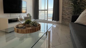 Un salon moderne doté d’un téléviseur à écran plat, d’une table basse en verre, d’un canapé et d’une grande fenêtre agrémentée de rideaux.