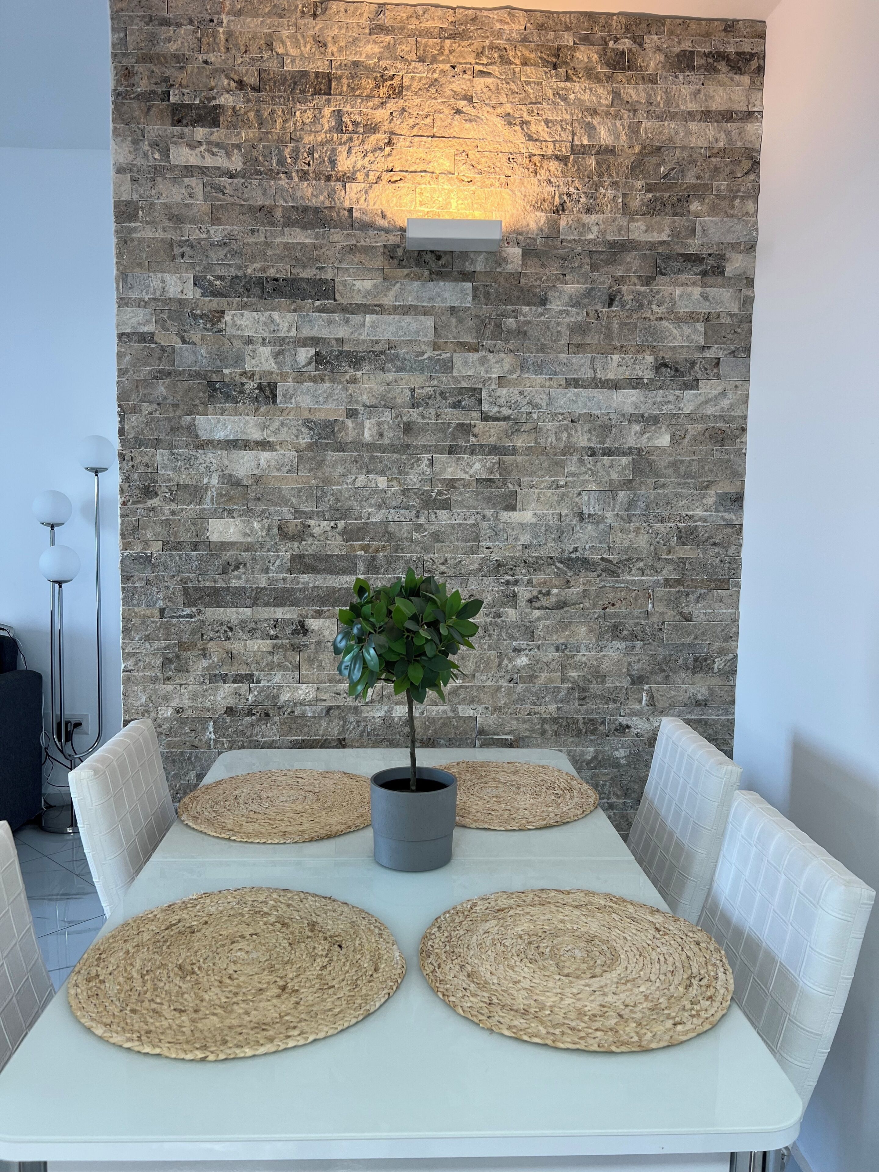 A dining table with four woven placemats, a potted plant, and a stone wall with a light fixture.