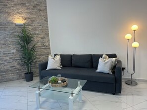 Un salon moderne avec un canapé gris foncé, deux coussins blancs, une table basse en verre agrémentée d’un plateau décoratif, un grand lampadaire avec trois abat-jour sphériques et une grande plante en pot.
