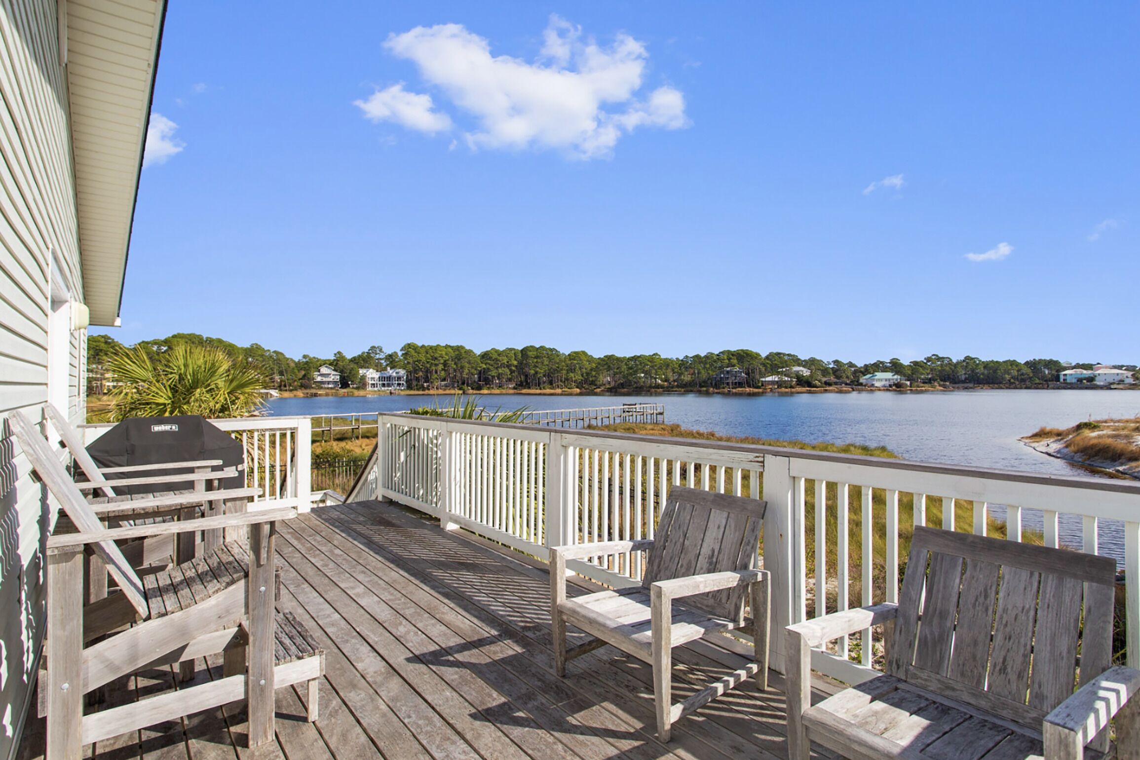 Eine Holzterrasse mit zwei Adirondack-Stühlen, einem Grill und Blick auf einen See mit Bäumen und Gebäuden im Hintergrund.