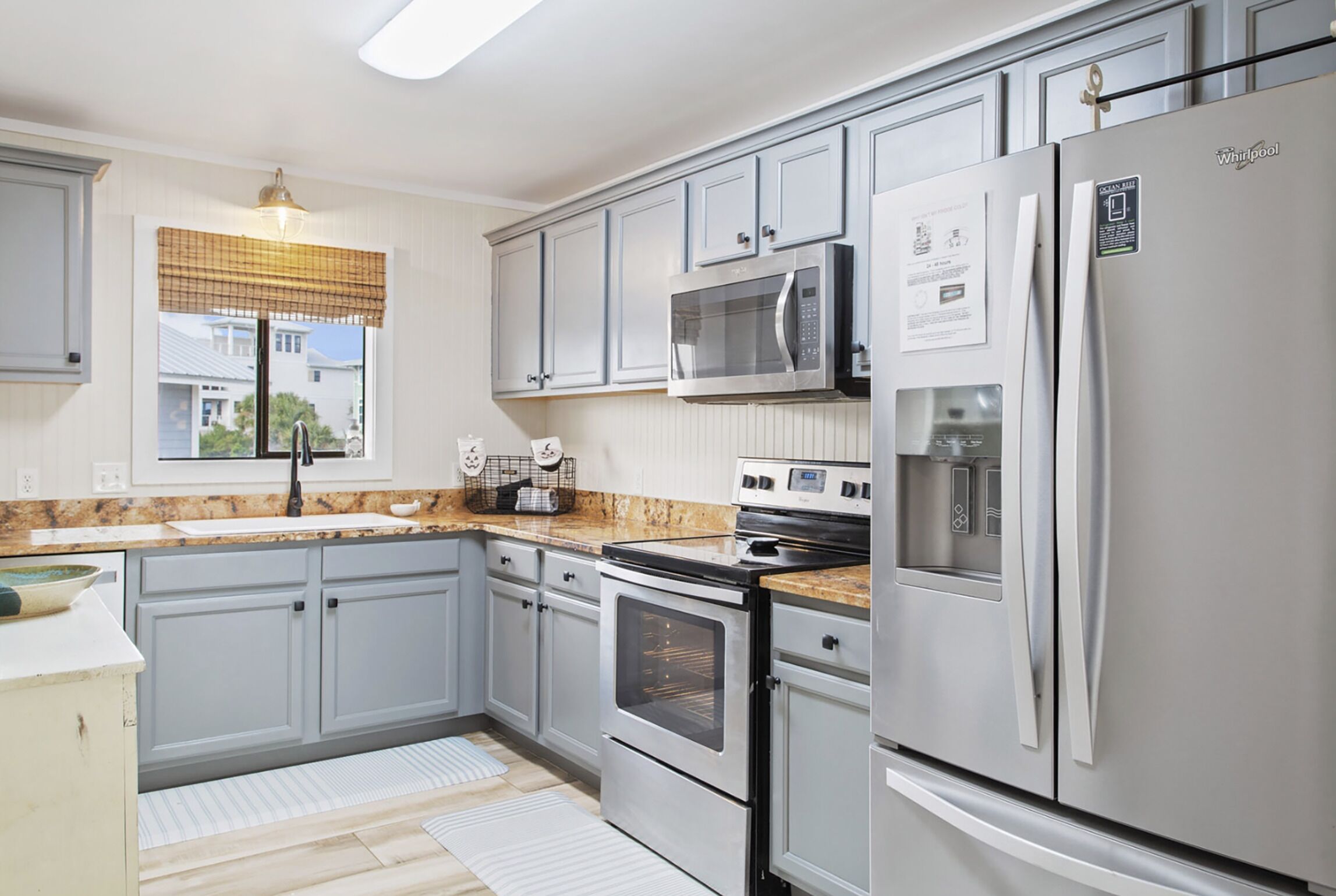 A kitchen with grey cabinets, a stainless steel refrigerator, a microwave, an oven, and a window with blinds.