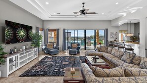 A spacious living room with a large sofa, a coffee table, and a TV. There is a ceiling fan, a large window with a view, and a dining area with chairs.