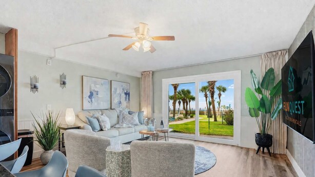A modern living room with a sofa, chairs, a coffee table, a ceiling fan, and a large window with a view of palm trees. - The Summit 131 - Ground Floor Beachfront! Walk to Sand! (Panama City Beach)