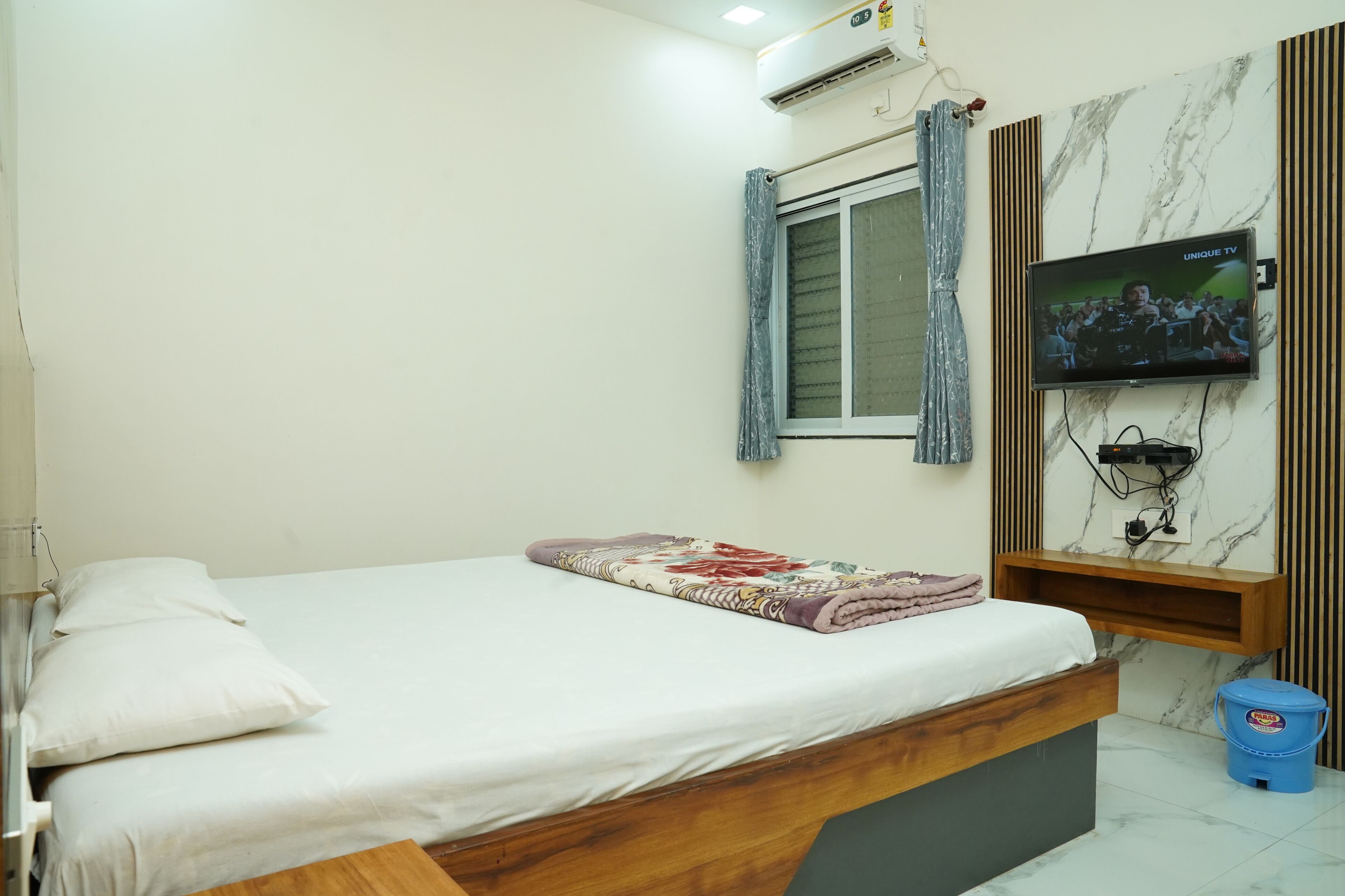 Un dormitorio con cama, televisor montado en la pared, ventana con cortinas y una pequeña estantería de madera.