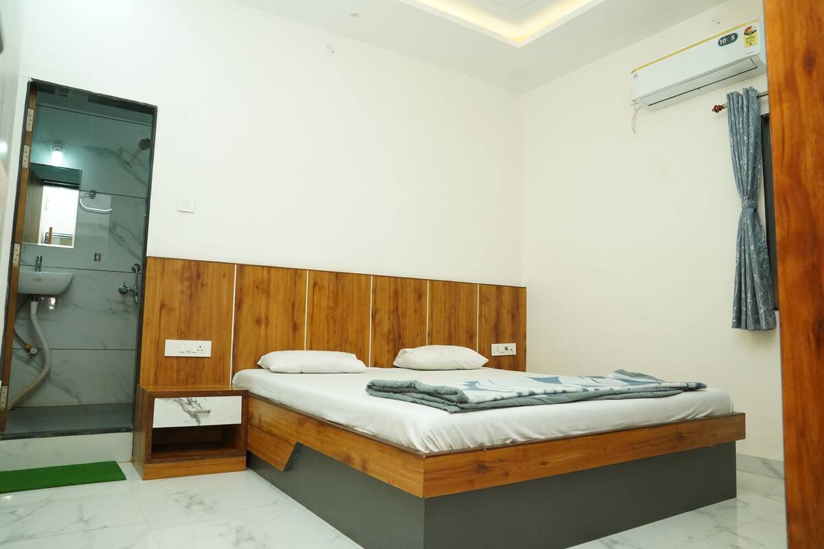 A bedroom with a wooden headboard, a bed with white bedding, a small bedside table, and a wall-mounted air conditioning unit.