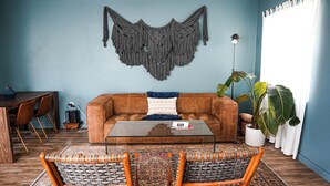 Uma sala de estar com um sofá marrom, duas cadeiras de vime, uma mesa de centro de vidro, um painel de macramé, uma planta em vaso e uma área de jantar com mesa e cadeiras de madeira.