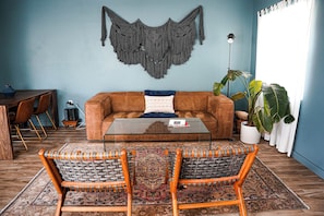 A living room with a brown sofa, two wicker chairs, a glass coffee table, a macrame wall hanging, a potted plant, and a dining area with a wooden table and chairs. - Cozy Villa Near Clearwater Beach with Patio & Hammock (Clearwater)