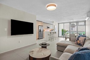 A modern living room with a flat-screen TV, a round coffee table, a sofa with cushions, and a dining area with chairs. - Brisanova | Modern Poolside Retreat - Top Location (Carolina)