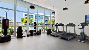 Un gimnasio con cintas de correr, pesas y balones de ejercicio, amplias ventanas con vistas a edificios y palmeras.