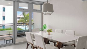 Un comedor moderno con una mesa blanca, sillas grises, una lámpara colgante y vistas a una piscina y edificios a través de amplios ventanales.
