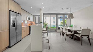 Una cocina moderna con electrodomésticos de acero inoxidable, un comedor con mesa y sillas blancas, y amplias ventanas que ofrecen vistas a zonas verdes.