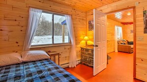 Ein gemütliches Blockhauszimmer mit Bett, Fenster mit Vorhängen, Lampe und einer Tür zu einem weiteren Raum.