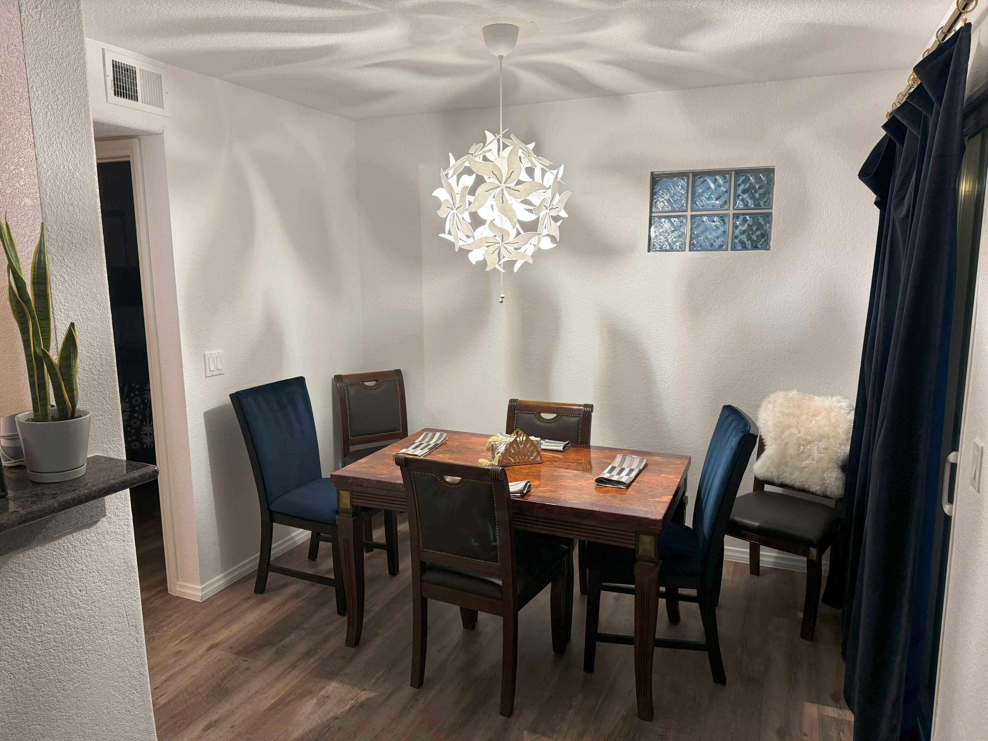 Un coin repas comprenant une table en bois, six chaises, un luminaire suspendu, une plante en pot et un élément décoratif mural.