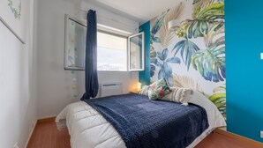 Ein Schlafzimmer mit Bett, einem Fenster mit Vorhängen, einer Wand mit einem tropischen Blatt-Wandbild und einem Nachttisch mit Lampe.