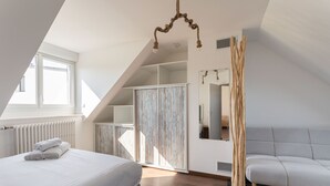 Quarto com cama, sofá cinzento, guarda-roupa de madeira, espelho e janela.