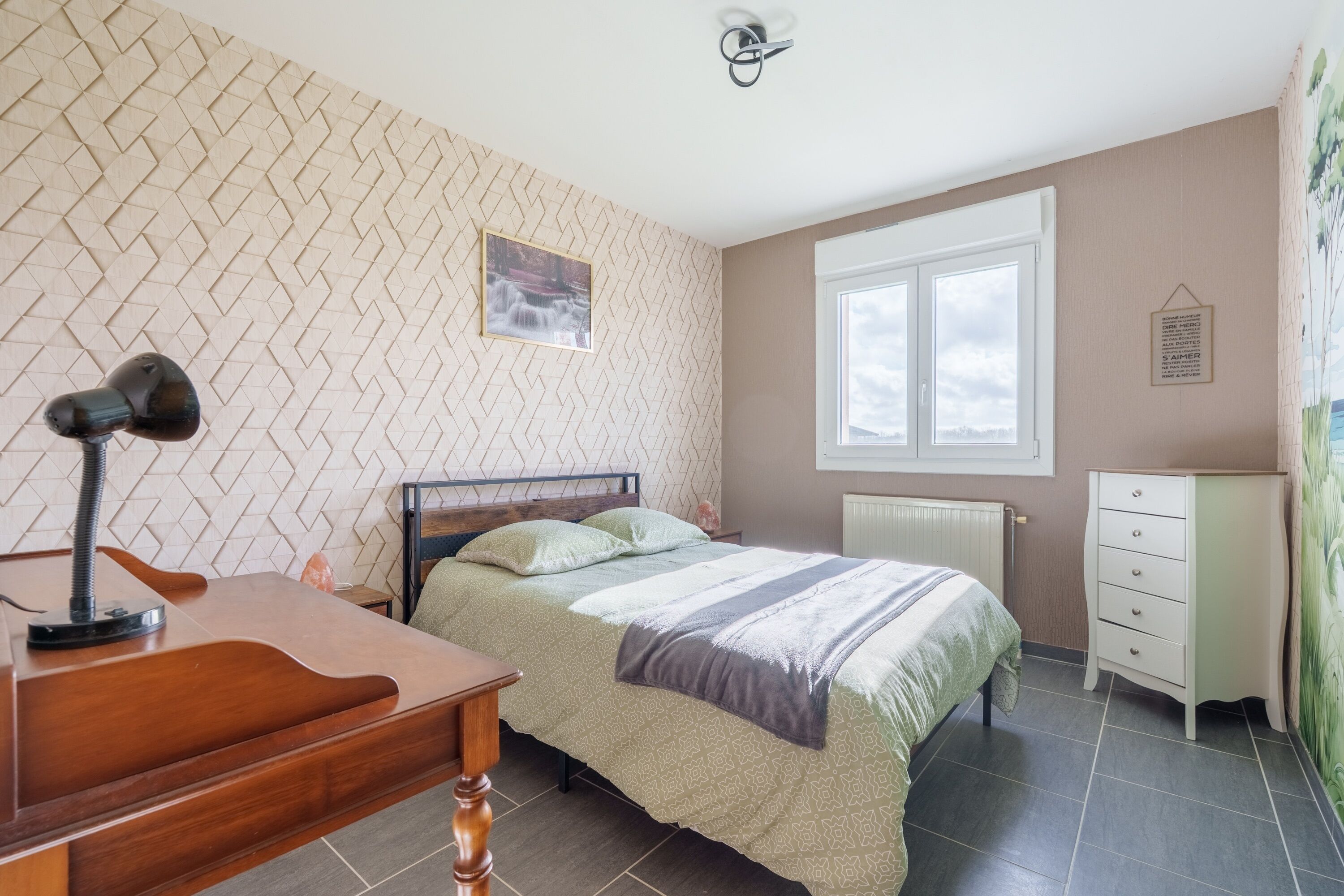 A bedroom with a bed, a desk, a chair, a radiator, a window, and a wall-mounted picture.