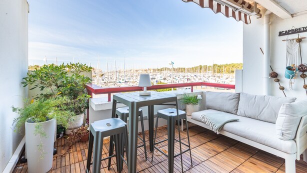 Ein Balkon mit einer weißen Couch, einem Tisch, Stühlen, Blumentöpfen und Blick auf einen Yachthafen.