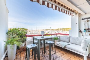 Un balcón con un sofá blanco, una mesa y sillas, plantas en macetas y vistas a un puerto deportivo.