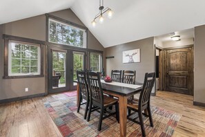 A dining area with a large table, chairs, a chandelier, and a view of trees through large windows. - 2 Bedroom Cabin with Fireplace (West Glacier)