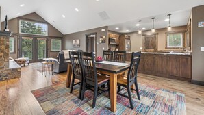 A spacious dining area with a wooden table and chairs, a kitchen with wooden cabinets, and large windows allowing natural light. - 2 Bedroom Cabin with Fireplace (West Glacier)