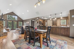 A spacious dining area with a wooden table and chairs, a kitchen with wooden cabinets, and large windows allowing natural light. - 2 Bedroom Cabin with Fireplace (West Glacier)