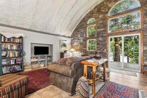 A cozy living room with a stone wall, a large window, a fireplace, and a bookshelf. - Two Bedroom Quonset Hut on Spoon Lake (Columbia Falls)