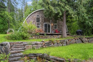 A stone house with a curved roof, surrounded by a stone wall and a grassy area. - Two Bedroom Quonset Hut on Spoon Lake (Columbia Falls)