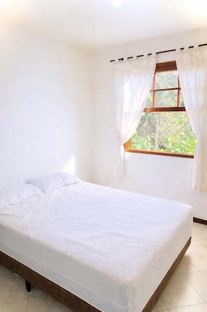 A bedroom with a single bed, a window with curtains, and a view of greenery outside.