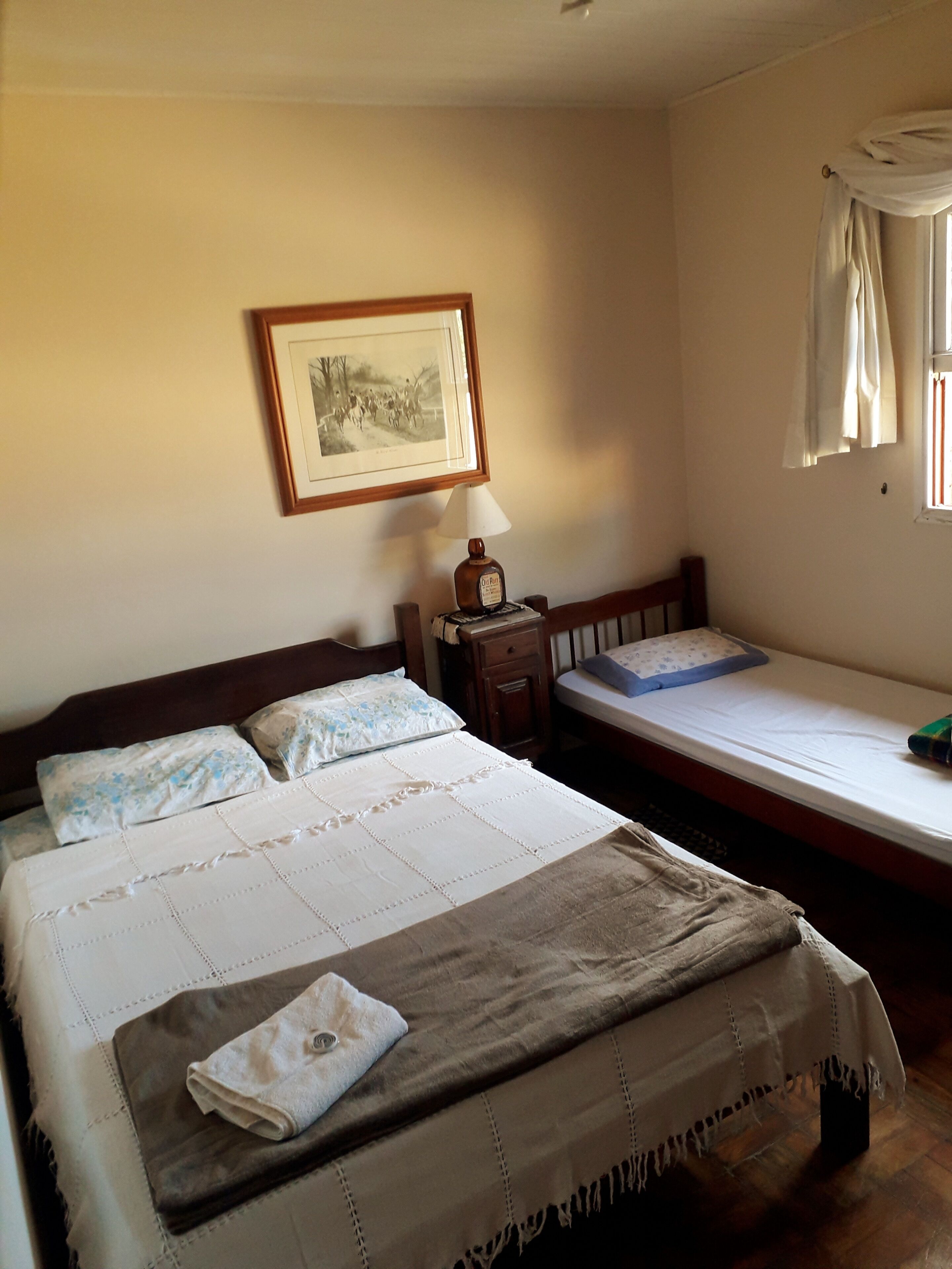 Ein Schlafzimmer mit zwei Betten, einem Nachttisch, einer Lampe, einem gerahmten Bild an der Wand und einem Fenster mit Vorhängen.