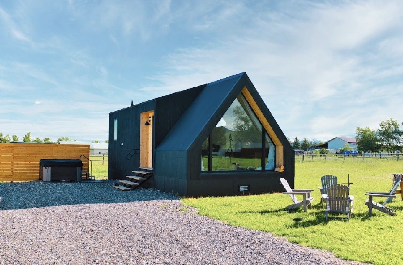 A modern, triangular-shaped cabin with a large window, a wooden door, and a gravel driveway.