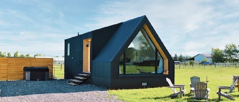 A modern, triangular-shaped cabin with a large window, a wooden door, and a gravel driveway.