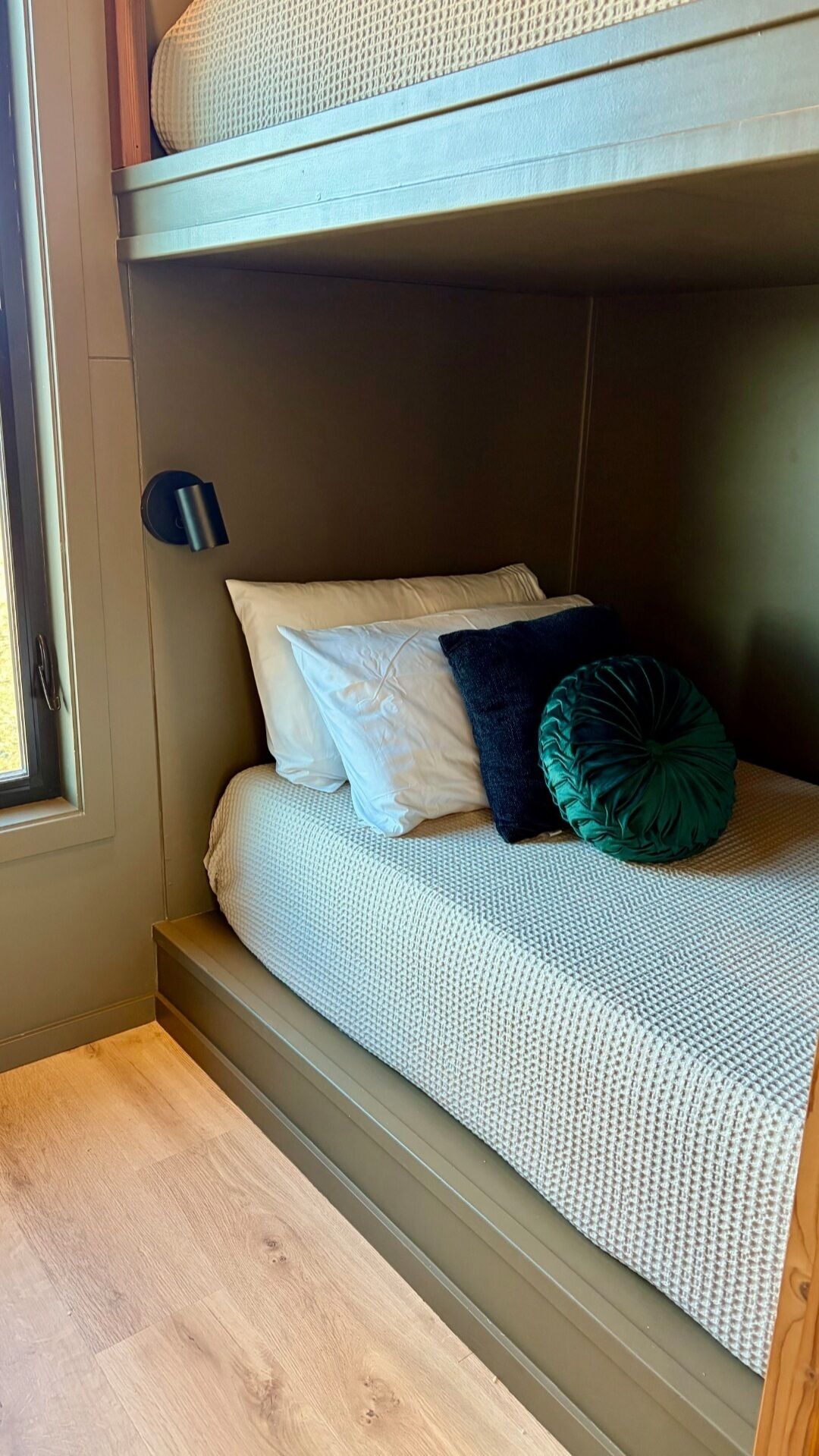 Un letto a castello con una finestra, una lampada fissata al muro e un cuscino verde.