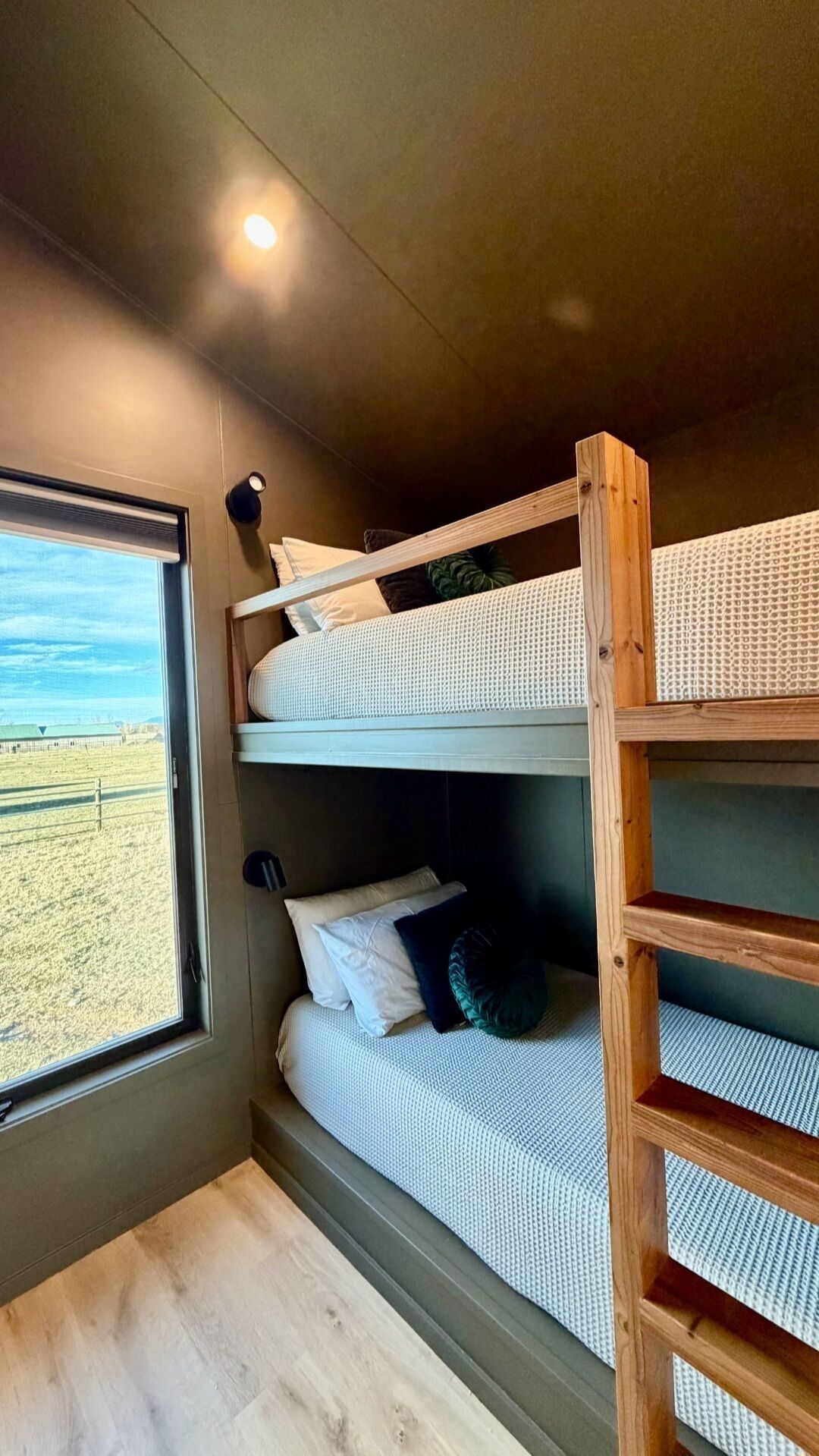 Un letto a castello con una scala in legno, una finestra vista su un campo e un piccolo ripiano.