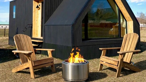 A modern cabin with a triangular roof, two wooden chairs, and a metal fire pit. - Cozy Luxe Cabin w/Hot Tub, Fire Pit & Stunning Views (Bozeman)