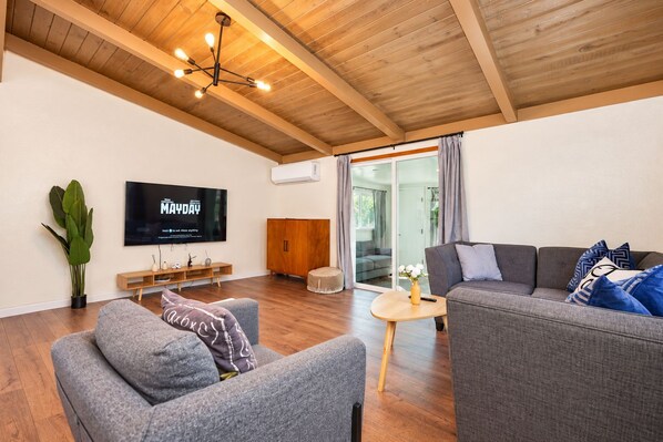 Ein modernes Wohnzimmer mit einem Flachbildfernseher, grauen Sofas, Holzböden und einer Decke mit freiliegenden Balken.