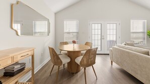 A modern living room with a round wooden table, wicker chairs, a beige sofa, a large mirror, and a wooden desk.