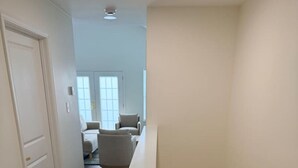 Un corridoio con una scala in legno, una porta bianca e vista su una stanza con un divano bianco e una finestra.