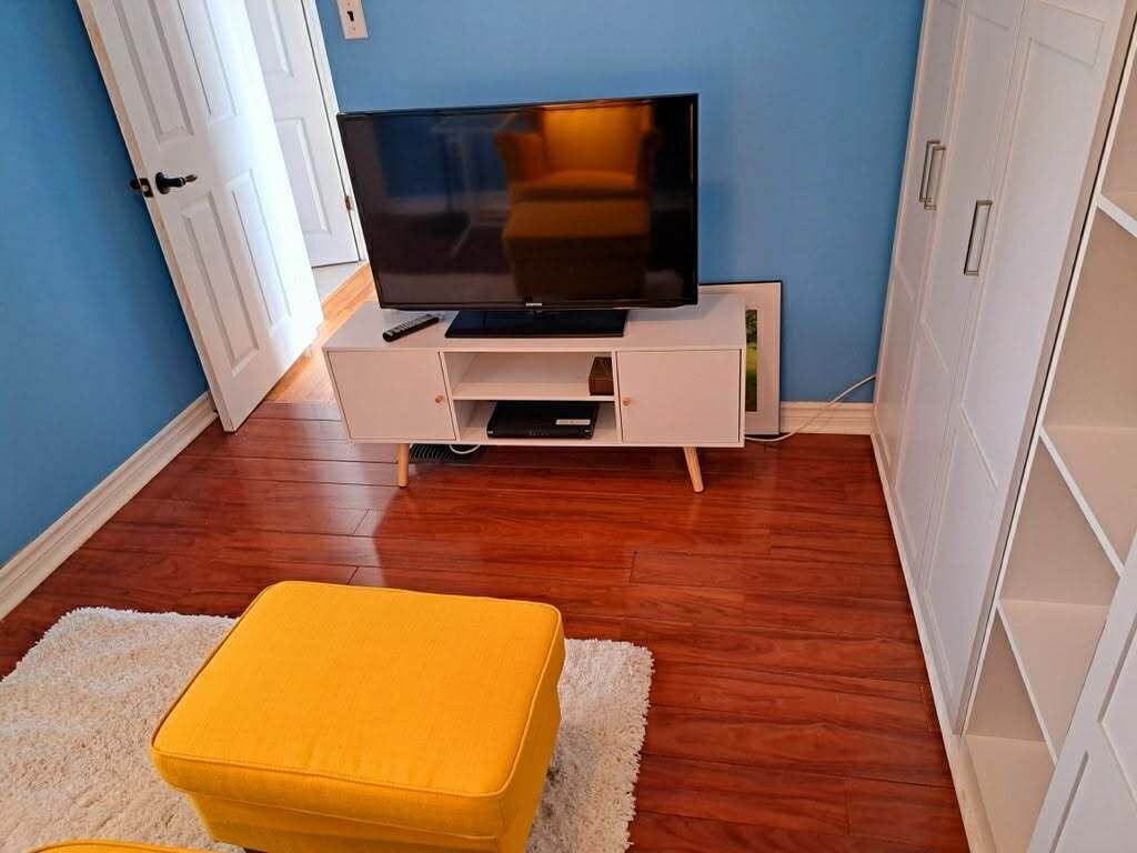 A room with a television on a white stand, a yellow ottoman, wooden flooring, and a blue wall.