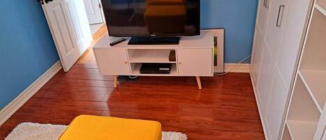 A room with a television on a white stand, a yellow ottoman, wooden flooring, and a blue wall.