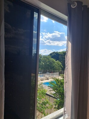 A view through a window with curtains, featuring a swimming pool, outdoor furniture, and a fenced area with greenery. - Apartamento com 2 quartos, a 15 minutos da praia Barra da Tijuca,  RJ, Brasil (Rio de Janeiro)