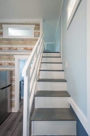 Una escalera con acabado de madera, barandas blancas y una zona de cocina con pared de madera rústica y un refrigerador.