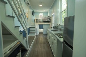 A narrow, multi-level kitchen with a bunk bed, wooden flooring, and a large window. - Harbor Blue (Anderson)