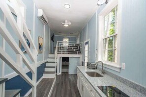 A narrow hallway with a staircase, a kitchen area with a sink, and a window with a view of greenery. - Harbor Blue (Anderson)