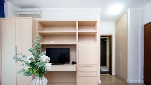 Quarto moderno com televisão sobre suporte de madeira, uma mesa branca com um vaso e piso de madeira.