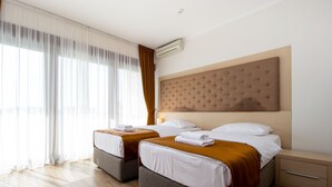 Quarto de hotel com duas camas, uma janela ampla com cortinas leves, ar condicionado e piso de madeira.