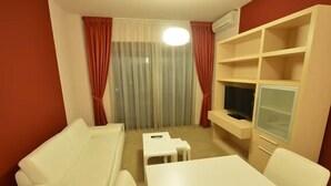 A living room with a white sofa, a small white table, a TV on a stand, and a window with red curtains. - Hotel Princ-One Bedroom Suite with Double Bed 102 3 (Budva)