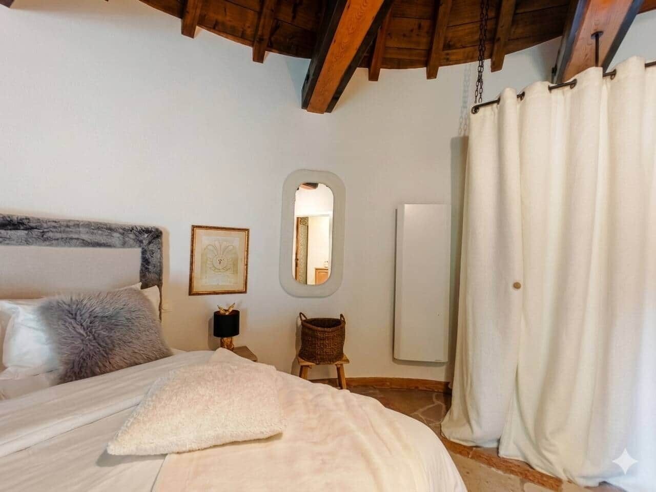 A bedroom with a bed, a nightstand, a mirror, a wicker chair, and a curtain.